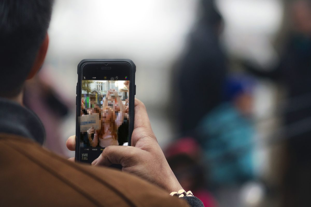 Une personne vue de dos filme avec son téléphone ; à l’écran, une manifestation où les visages sont pixelisés